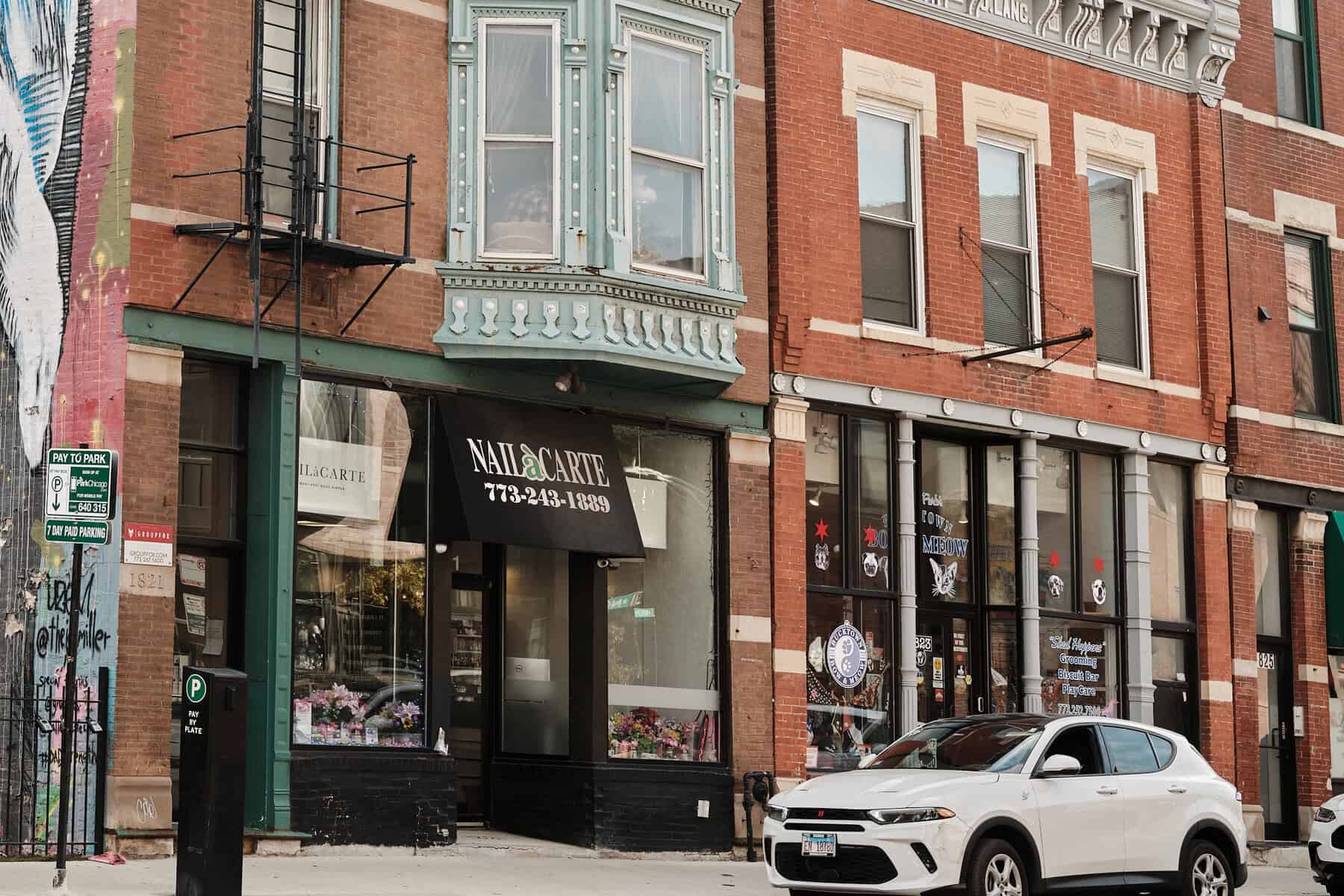Colorful nail salon storefront with urban brick building exterior and parked white SUV in front.